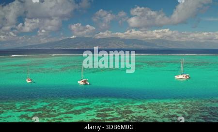 Die Landschaft der Insel Moorea bietet einen Blick aus der Vogelperspektive auf das türkisfarbene und blaue Lagunenwasser, mit Segelbooten, die über einem flachen Korallenriff vertäut sind, und eine ferne Insel, die eine ruhige tropische Szene schafft Stockfoto