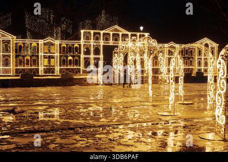 Festlicher Gehweg-Tunnel aus goldenen Weihnachtslichtbögen. Urbane Nachtkulisse mit leuchtenden Dekorationen, die sich auf nassem Boden spiegeln. Stockfoto
