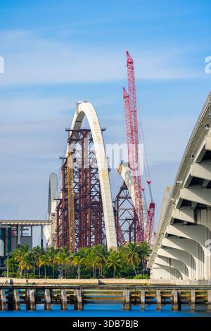 Neue Brücke im Bau in Downtown Miami. Ansicht der Bögen und Krane 2025. Lange Belichtung für glattes Wasser und Himmel Stockfoto