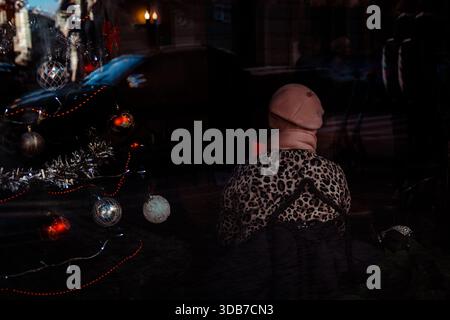 KIEW, Ukraine 13. Dezember 2025 Eine Frau mit Leopardenmuster und rosa Hut sitzt in einem Stadtcafé neben einem hell erleuchteten Weihnachtsbaum. Die Stockfoto