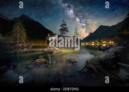 Magical blue scene at a lake in the night, with a man standing under the Milky Way on a rock and holding a light over the calm water Stockfoto