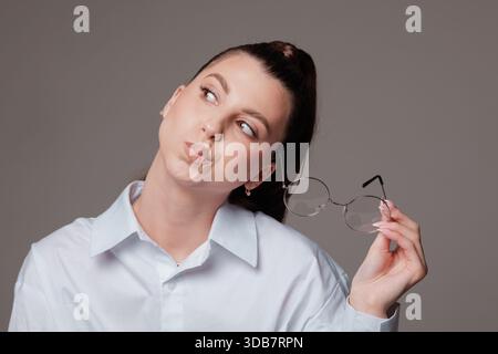 Lächelnde junge Frau mit Zahnspangen, die eine runde Brille halten, selbstbewusster und glücklicher Ausdruck, stilvoll in einem weißen Hemd, modernes Porträt auf grauem Hintergrund. Stockfoto