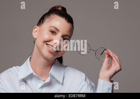 Lächelnde junge Frau mit Zahnspangen, die eine runde Brille halten, selbstbewusster und glücklicher Ausdruck, stilvoll in einem weißen Hemd, modernes Porträt auf grauem Hintergrund. Stockfoto