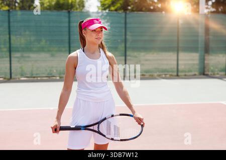 Junge Tennisspielerin in weißer Sportbekleidung mit Schläger, die im Sonnenlicht auf dem Platz steht und selbstbewusst lächelt. Stockfoto