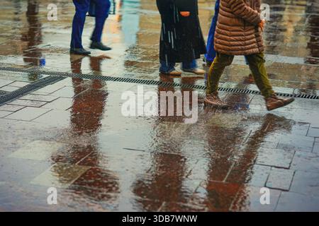 Menschen, die bei Regen auf nassen Straßen laufen, nur durch ihre Reflexionen und Beine in Pfützen sichtbar. Konzept des Stadtrhythmus, des Stadtwetters, der Saison Stockfoto