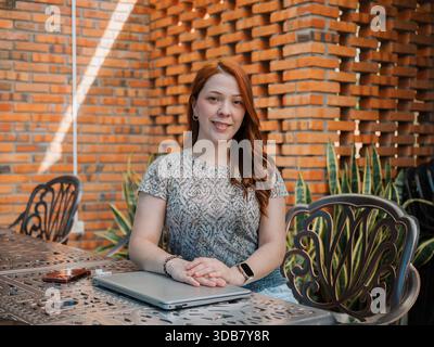 Lächelnde Frau sitzt an einem Tisch im Freien mit einem Laptop vor sich. Konzept für Fernarbeit, Studium oder Online-Meetings. Stockfoto