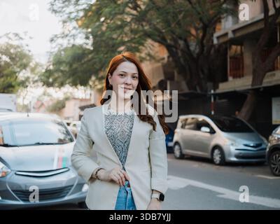 Eine junge Geschäftsfrau steht auf einer Straße mit Autos im Hintergrund. Professionelles Selbstvertrauen und urbanes Lifestyle-Konzept. Stockfoto