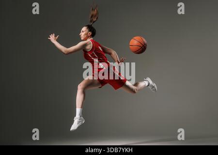 Junge Basketballspielerin, die mitten in der Luft mit Ball hüpft, voller Energie und Kraft, isoliert auf einem grauen Hintergrund im Studiolicht. Stockfoto