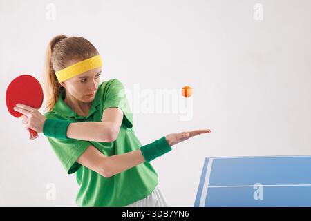 Eine konzentrierte Tischtennisspielerin bereitet sich auf das Servieren vor und wirft beim Spielbeginn einen orangen Ball in die Nähe des Paddels. Stockfoto