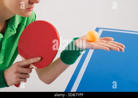 Nahaufnahme einer Athletin mit rotem Paddle und orangefarbenem Tischtennisball, fokussiert vor dem Servieren beim Tischtennis. Stockfoto