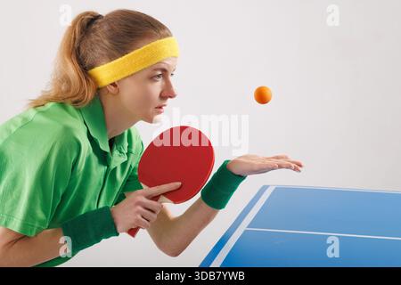Konzentrierte Athletin im grünen Polo und Stirnband bereitet sich darauf vor, orangen Tischtennisball beim Tischtennisspiel zu servieren. Stockfoto