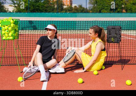 Zwei junge Tennisspielerinnen ruhen sich auf dem Platz am Netz aus und unterhalten sich mit Schlägern und Tennisbällen. Stockfoto