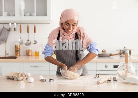 Frau, die Teig in einer Küche mit einem Lächeln zubereitet, während sie einen Hijab und eine Schürze während einer Kochsitzung trägt Stockfoto