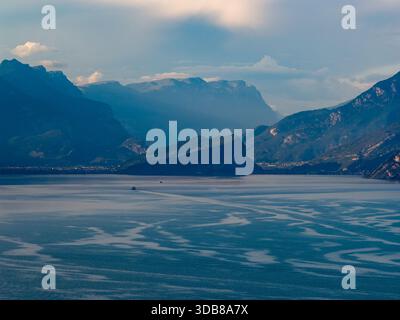 Blick aus der Vogelperspektive über den Gardasee in Richtung Malcesine, eingerahmt von überlagerten Alpenkämmen unter trübem Sommerlicht am späten Nachmittag. Stockfoto