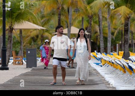 Ein paar Spaziergänge Hand in Hand entlang einer palmengesäumten Promenade in Harvest Caye, Belize, mit Reihen von Strandliegen und Sonnenschirmen tropischen Kreuzfahrtanleger Stockfoto
