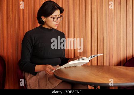 Eine junge Frau, die an einem Café-Tisch sitzt, Brille und einen schwarzen gerippten Rollkragen trägt, intensiv ein Buch mit Konzentration im Gesicht liest und genießt Stockfoto