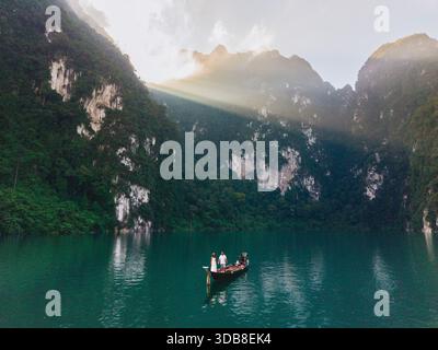 Erleben Sie das ruhige Wasser des Cheow Lan Lake in Khao Sok, Thailand bei Sonnenaufgang. Ein Paar in einem Langboot gleitet sanft über den See, während die Sonne goldene Strahlen auf die hohen Klippen wirft. Stockfoto