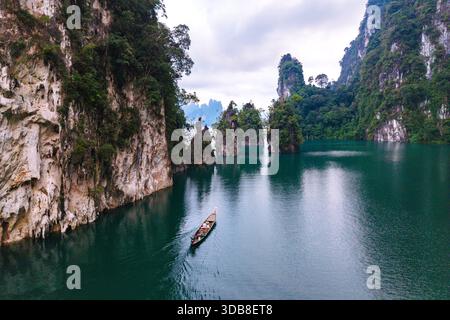 Erleben Sie das ruhige Wasser des Cheow Lan Lake in Khao Sok, Thailand. Ein langes Boot gleitet durch smaragdgrüne Gewässer, umgeben von majestätischen Kalksteinklippen und üppigem Grün, was einen friedlichen Rückzugsort schafft. Stockfoto