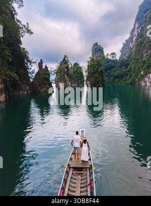 Erleben Sie die ruhige Atmosphäre des Cheow Lan Lake, umgeben von üppigem Grün und hoch aufragenden Kalksteinklippen. Ein Paar vor einem Langboot in Khao Sok Thailand Stockfoto