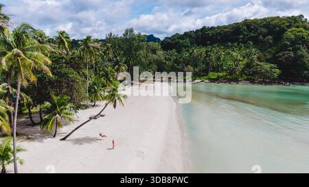 Weicher weißer Sand trifft auf kristallklares Wasser auf Koh Kood Island in Thailand. Üppige Palmen schwingen sanft unter einem hellen Himmel. Ein paar Männer und eine Frau auf einer Schaukel unter einer Palme am Strand Stockfoto