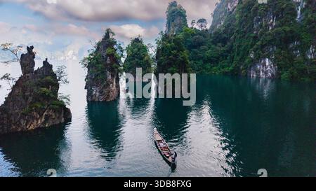 Erkunden Sie das ruhige Wasser des Cheow Lan Lake in Khao Sok, Thailand. Hoch aufragende Kalksteinklippen bieten eine majestätische Kulisse, während Longtail-Boote durch das ruhige smaragdgrüne Wasser unter einem bewölkten Himmel gleiten. Stockfoto