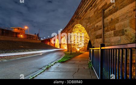 Stone Arch Bridge features illuminated arches at night in Minneapolis. Long exposure captures light trails on West River Parkway under a cloudy sky Stockfoto