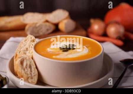 Warme und gemütliche Herbstsuppenszene mit Kürbissuppe garniert mit Sahne und Samen, ergänzt durch rustikale Brotscheiben in einer einladenden Küche A Stockfoto