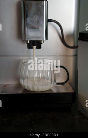 Automatische Kaffeemaschine, die Milch in einen klaren, gerippten Glasbecher mit schwarzem Griff in moderner Kücheneinrichtung abgibt, Rohr, Glas, geschwungene, verschüttete plasti Stockfoto