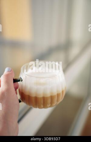 Eine Hand hält eine Glasschale aus geschichtetem Latte Macchiato mit cremigem Schaum in weichem natürlichem Licht, reflektiert vor einem verschwommenen Hintergrund, wodurch ein gemütliches A entsteht Stockfoto