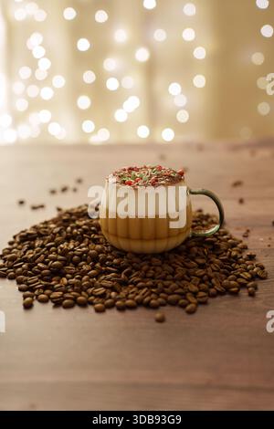 Festlicher Cappuccino in Glasbecher mit bunten Streuseln und Schaum, umgeben von Kaffeebohnen auf rustikaler Holzoberfläche mit weichem Bokeh-Licht-bac Stockfoto