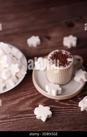 Eine gemütliche Szene mit einer Tasse heißer Schokolade mit Schokoladenstreuseln auf einem rustikalen Holztisch, umgeben von schneeflockenförmigen Marshmallows auf einer Cerami Stockfoto