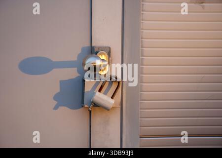 Nahaufnahme einer Metalltür mit einem glänzenden runden Türknauf mit einer angebrachten Metallplatte, die lange Schatten auf eine strukturierte Oberfläche wirft, gesichert mit Stockfoto