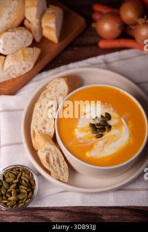 Warme und einladende Herbstmahlzeit mit Kürbissuppe garniert mit Sahne und Kürbiskernen, begleitet von rustikalem Brot und frischem Gemüse Stockfoto