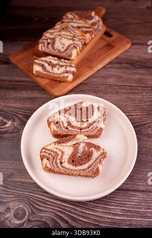 Marmorkuchen auf einem Holzbrett und weißer Teller mit üppigen Schokoladen- und Vanillewirbeln auf einer rustikalen Holztischoberfläche, künstlerisch, i Stockfoto