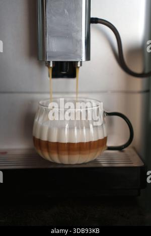 Nahaufnahme einer Espressomaschine, die reichhaltigen Schichtkaffee mit aufgeschäumter Milch in einer Glasschale auf der modernen Küchenarbeitsfläche zubrüht, verlockend, raffiniert, Stockfoto