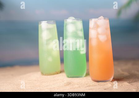 Drei lebhafte, mit Eis gefüllte tropische Getränke in hohen Gläsern am Sandstrand mit verschwommenem Meer und Palmblättern im Hintergrund, die an frischen Sommer erinnern Stockfoto