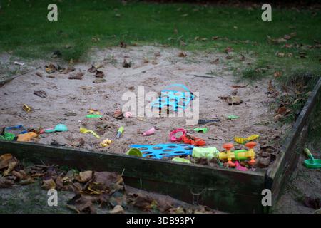 Verlassene Sandkiste mit bunten Spielzeugen inmitten gefallener Blätter an einem ruhigen bewölkten Tag in einer grasbewachsenen Umgebung im Freien, Abenteuer, lässig, isoliert, unordentlich, rev Stockfoto