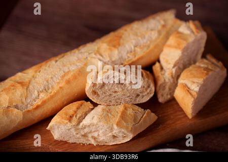 Nahaufnahme eines frisch gebackenen Baguettes und in Scheiben geschnittener Brotstücke auf einem Holzbrett mit luftiger Textur und goldener Kruste vor einem warmen, gemütlichen Background Stockfoto