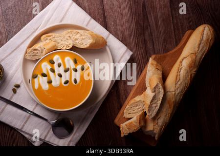 Blick von oben auf herbstliche Suppe garniert mit Samen und Creme, begleitet von frischen Baguette-Scheiben auf rustikalem braunen Holztisch mit weicher warmer Feuchte Stockfoto