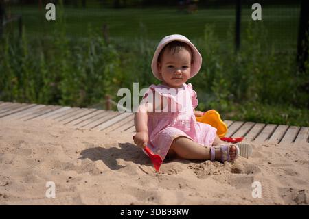 Ein charmantes Kleinkind in rosa Kleid und Sonnenhut genießt einen sonnigen Tag in der Sandbox, umgeben von üppigem Grün, mit farbenfrohen Sandspielzeugen in einer ruhigen Umgebung Stockfoto