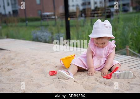Ein Kleinkind in rosa Kleid und Sonnenhut spielt friedlich mit roter Schaufel und gelbem Eimer in einem Sandkasten, umgeben von weichem Grün und einem ruhigen Stadtpark Stockfoto