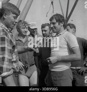 1970er Jahre UDSSR. Region Donezk. Bau strategischer 750-kV-Hochspannungsleitungen. Eine Gruppe junger Installateure und Ingenieure, die auf der Baustelle eine Pause machen. Die Männer stehen neben massiven Stahlkonstruktionen und einem Kranhaken, die wegen der Hitze in Sommerkleidung gekleidet sind. Sie rauchen Zigaretten, lachen und unterhalten sich lebhaft. Ein Dokumentarfilm, der die Atmosphäre der männlichen Freundschaft und den Arbeitsrhythmus der Energiebauer in der friedlichen Donbass-Steppe vor dem Krieg einfängt. Stockfoto