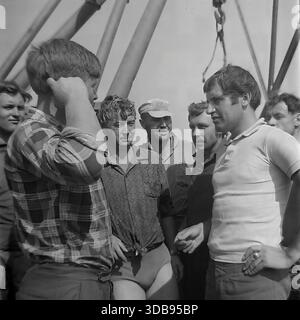 1970er Jahre UDSSR. Region Donezk. Bau strategischer 750-kV-Hochspannungsleitungen. Eine Gruppe junger Installateure und Ingenieure, die auf der Baustelle eine Pause machen. Die Männer stehen neben massiven Stahlkonstruktionen und einem Kranhaken, die wegen der Hitze in Sommerkleidung gekleidet sind. Sie rauchen Zigaretten, lachen und unterhalten sich lebhaft. Ein Dokumentarfilm, der die Atmosphäre der männlichen Freundschaft und den Arbeitsrhythmus der Energiebauer in der friedlichen Donbass-Steppe vor dem Krieg einfängt. Stockfoto