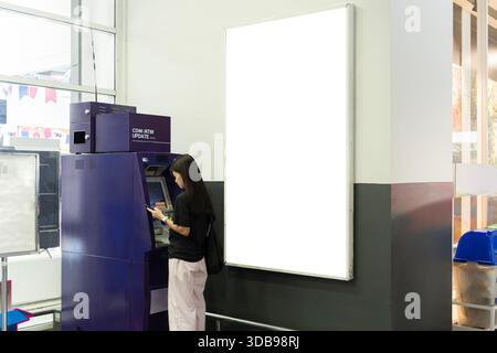 Frau, die einen Geldautomat in einem öffentlichen Gebäude mit großen leeren Werbetafeln benutzt, die Bankdienstleistungen, Finanztransaktionen, Bargeld repräsentieren Stockfoto