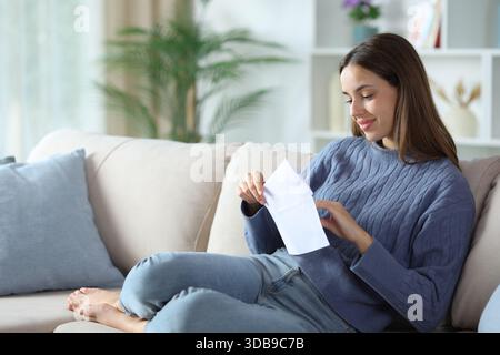 Frau in Blau, die einen Brief in einen Umschlag legt, sitzt zu Hause auf einem Sofa Stockfoto