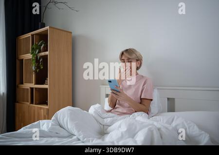 Verschlafene Frau mittleren Alters, die die Morgenzeit im Bett verzögert und soziale Medien auf dem Smartphone durchblättert. Stockfoto