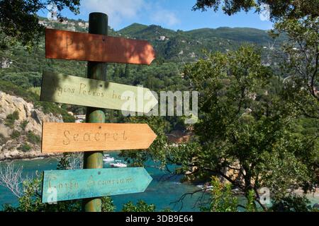 Farbenfrohe und rustikale hölzerne Wegweiser zu Zielen wie Agios Petros Beach Seacret und Korfu Aquarium mit einem Hintergrund aus grünen Hügeln und blauem Meer. Stockfoto