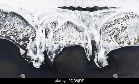 Aus der Vogelperspektive blickt der schroffe schwarze Sand auf eisweiße Weiten, ein Tanz aus Feuer und Eis, wo das Land das Meer umschließt, Hafnarfjordur, Hafnarfjardarkaups Stockfoto