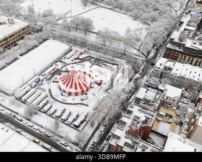 Aus der Vogelperspektive ein leuchtendes rot-weiß gestreiftes Zirkuszelt, eingebettet in eine schneebedeckte Stadt und einen Park, der einen starken Kontrast zum gedämpften Winter bildet Stockfoto