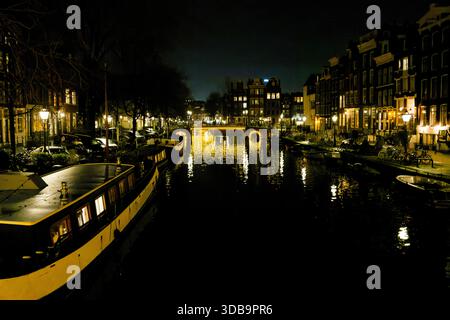 Das ruhige Wasser reflektiert das Leuchten der Straßenlaternen entlang eines malerischen Kanals in Amsterdam bei Nacht und zeigt bezaubernde Architektur und Boote, die entlang liegen Stockfoto